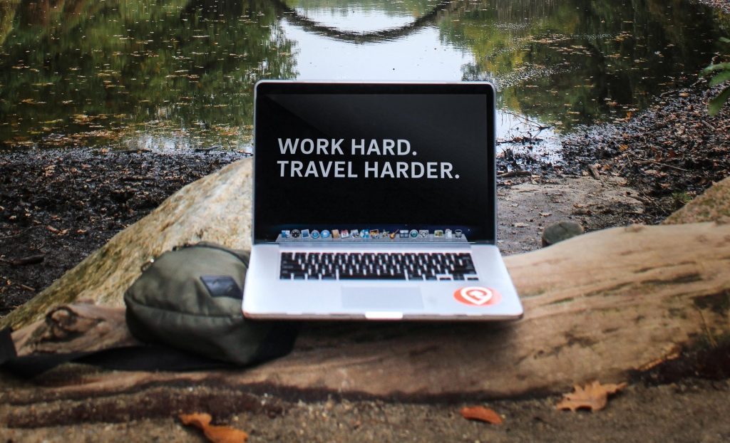MacBook on top of a log showing screen "Word Hard. Travel Harder"